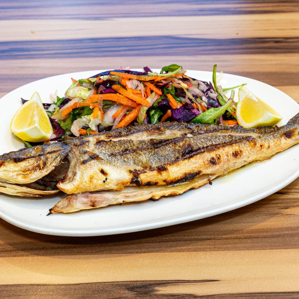 Grilled whole fish with a side of colorful salad and lemon wedges on a white plate.