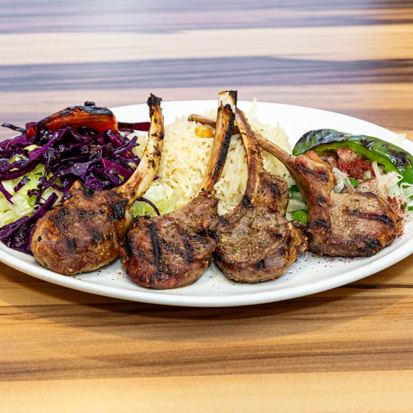 Four grilled lamb chops with rice, red cabbage salad, and grilled green pepper on a white plate.