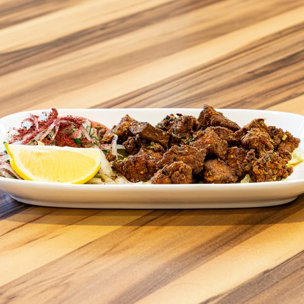 Turkish fried liver cubes with sliced onions, red spices, and a lemon wedge on a white plate.